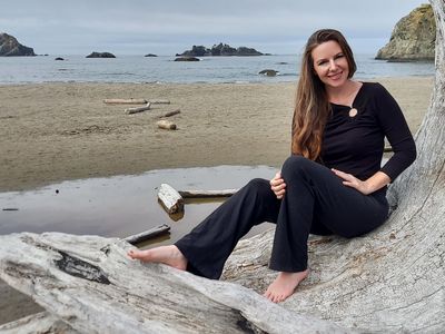 A woman sitting barefoot on a large driftwood log by the beach, smiling.