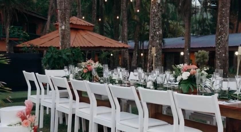 Mesa larga con sillas blancas y decoración floral al aire libre.