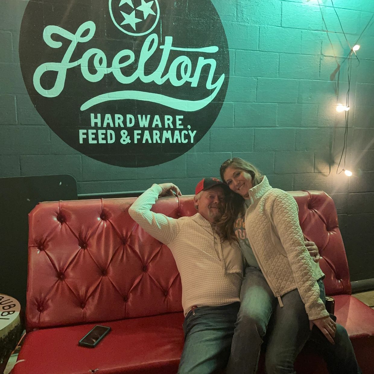 Couple smiling and relaxing on a red couch under a sign at Joelton Hardware.