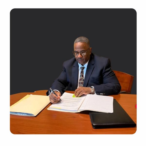 Man in suit signing documents at desk with folders and notebook.