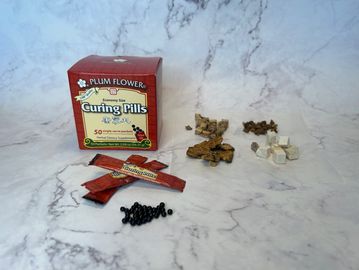 box of curing pill packets alongside small piles of raw herbs showing some of the ingredients