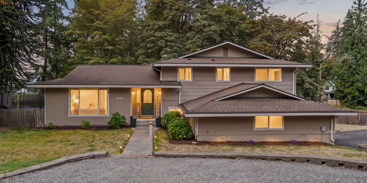 A modern two-story house with a lit interior and a gravel driveway at dusk.