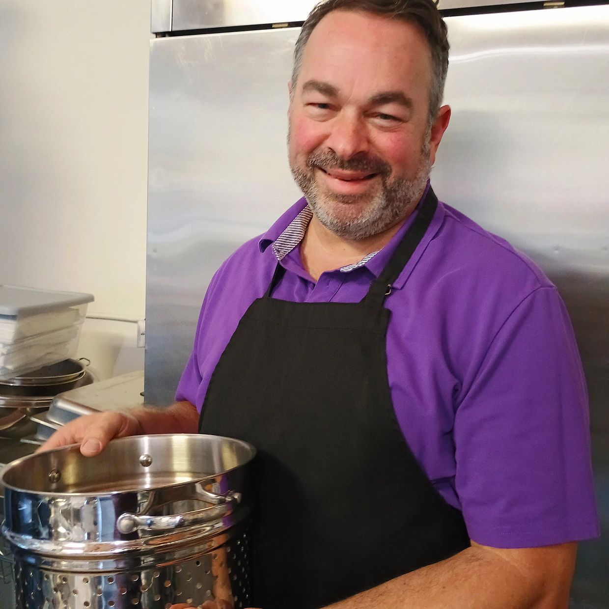 Smiling man in purple shirt and black apron holding a stainless steel pot.