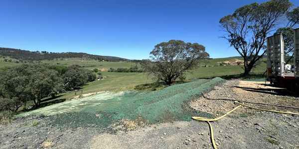 Rural landscape with green hills, scattered trees, and a truck on a gravel path under a clear blue sky.