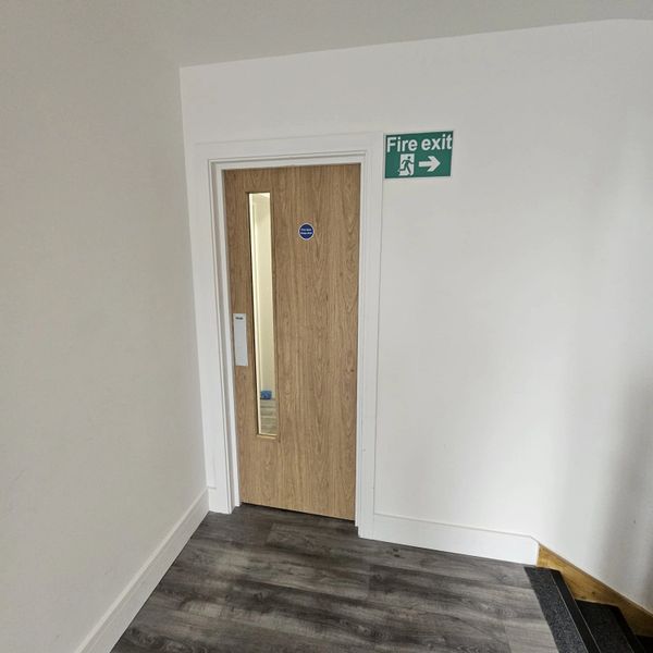 Wooden door with a fire exit sign above it in a white hallway.