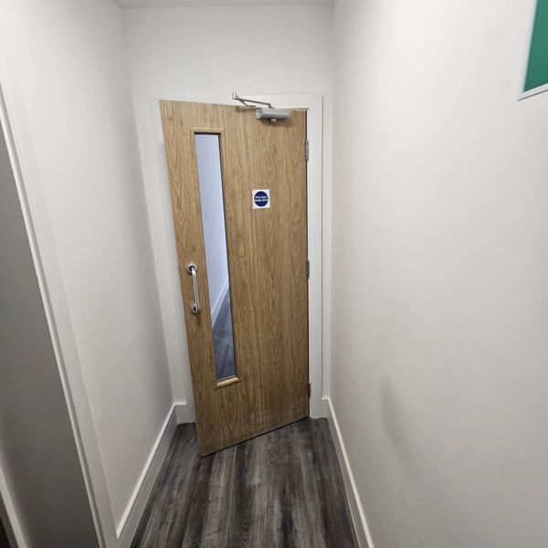 Wooden door with glass panel in a narrow hallway with white walls and gray flooring.
