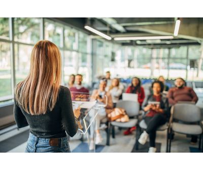 A woman is standing speaking to a group of people sitting in front of her. 