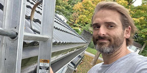 Man taking a selfie on a rooftop ladder with autumn trees in the background.