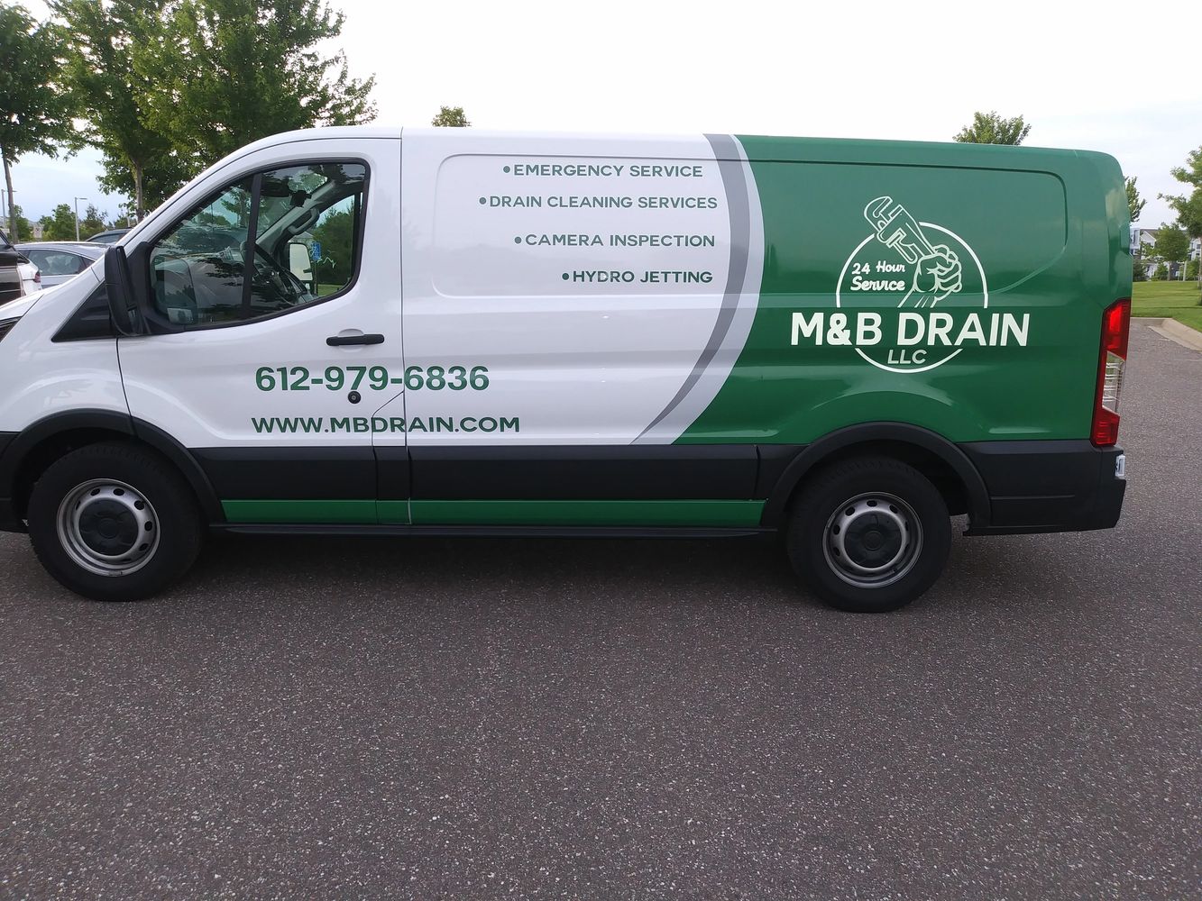 Side view of a white and green M&B Drain service van with contact details and services listed.