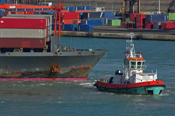 A tugboat guiding a large cargo ship near a busy port with containers.
