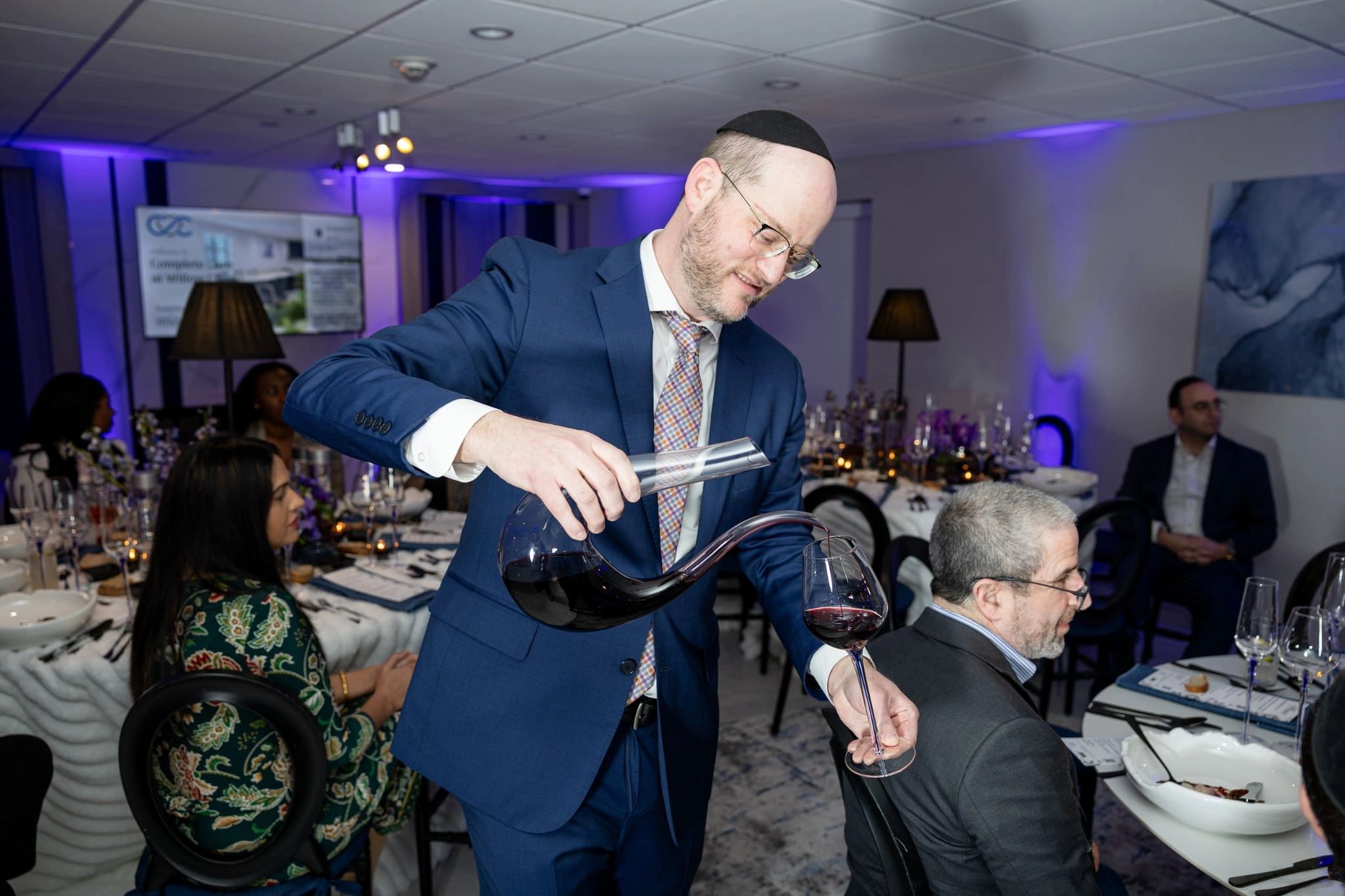 Man in a suit and kippah pouring red wine at a formal dinner.