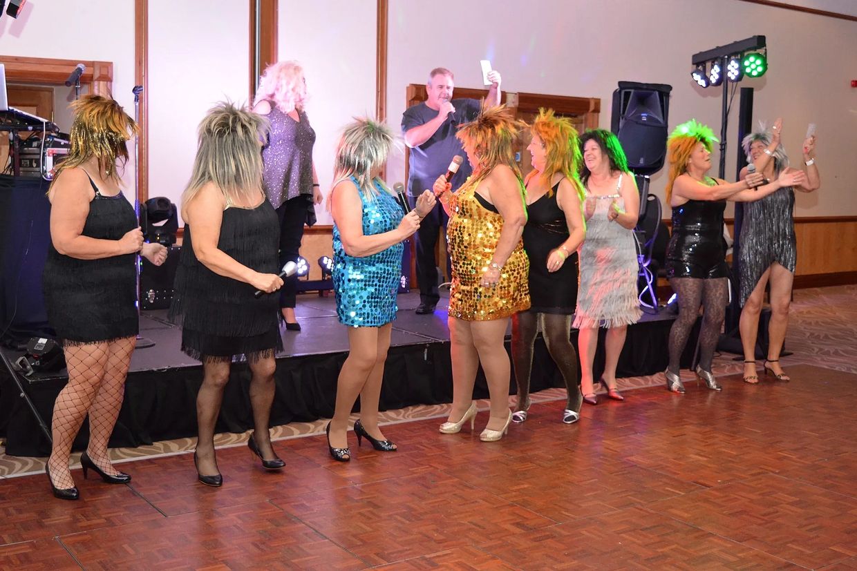 Women in sparkly dresses and wigs performing on stage with a man holding a microphone.