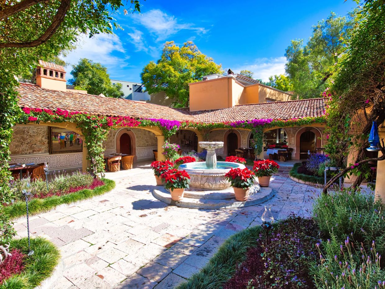 Sunny courtyard with a central fountain surrounded by potted red flowers and arched walkways.