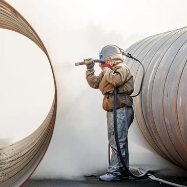 Worker sandblasting a large metal pipe wearing protective gear.