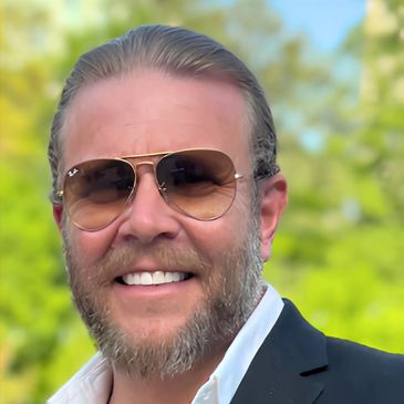 Smiling man with sunglasses and beard in a suit outdoors.