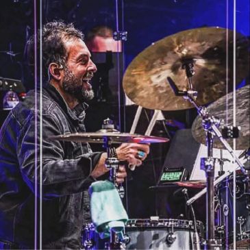 Drummer enthusiastically playing drums behind a transparent screen on stage.