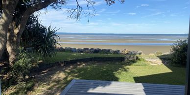 Beach view from a deck with trees and rocks under a clear blue sky.