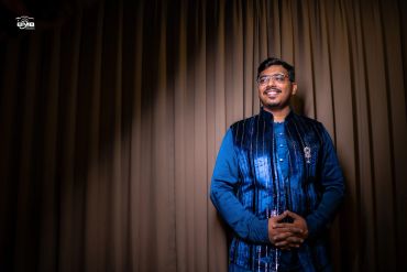 Smiling man in traditional blue attire standing by brown curtains.