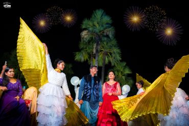 Couple surrounded by performers with golden wings under fireworks.