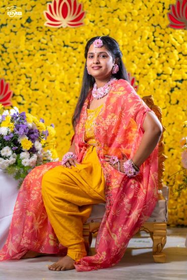 Woman in traditional attire with floral jewelry sitting on a chair.