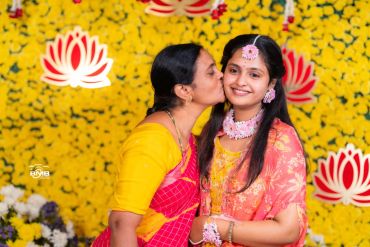 A woman kisses a young woman in traditional attire with floral jewelry.