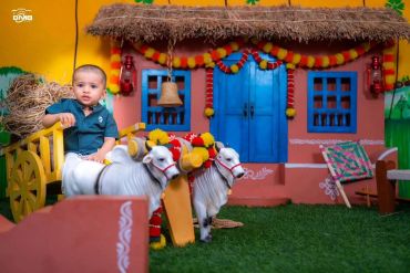 A toddler sitting on a decorative bullock cart in front of a colorful rustic house.