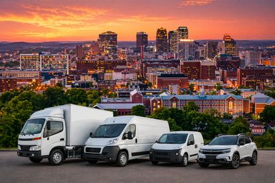A lineup of delivery vehicles with a city skyline at sunset.