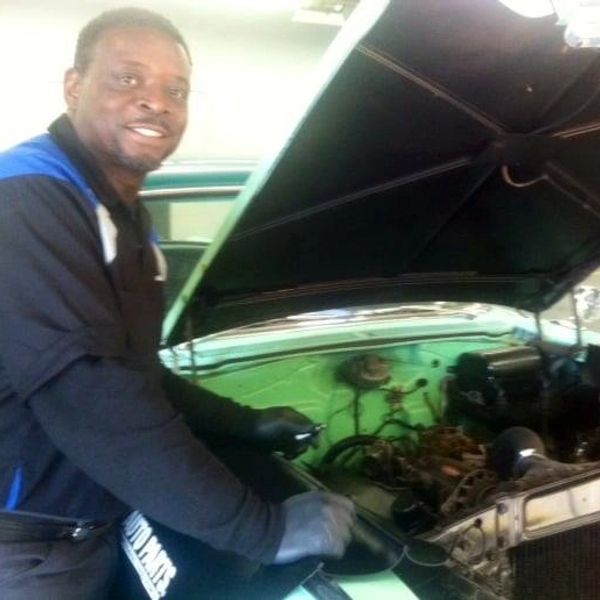 Mechanic smiling while working on a car engine with the hood open.