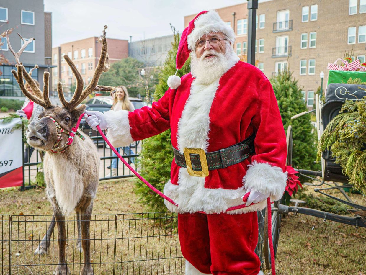 Santa Claus holding a reindeer on a leash outdoors.