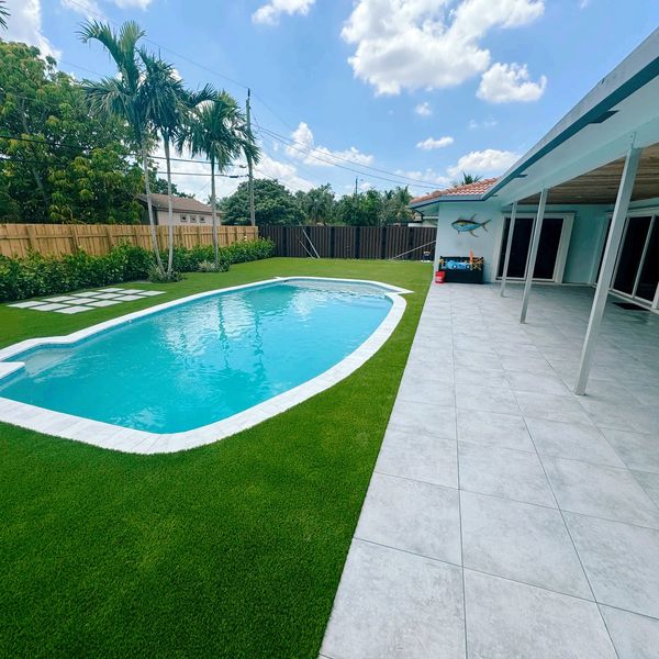 Backyard with a swimming pool, green lawn, and tiled patio under a partly cloudy sky.