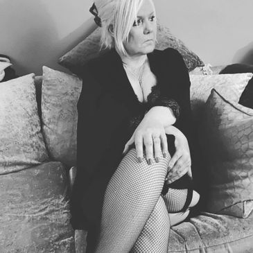 Woman in black outfit with fishnet stockings sitting on a couch, looking contemplative.
