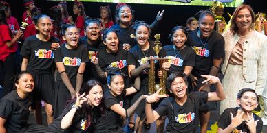 A joyful group of young girls celebrating with a trophy and adults on stage.
