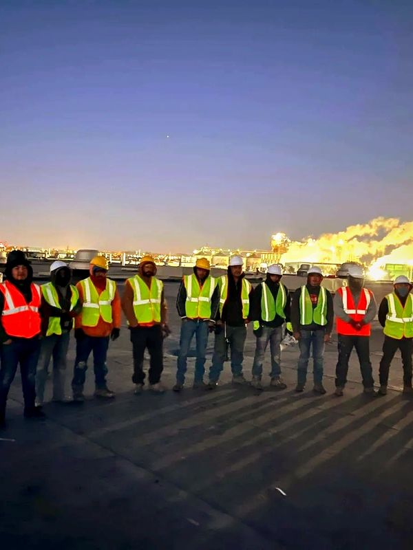 Trabajadores con chalecos reflectantes y cascos en un sitio industrial al atardecer.