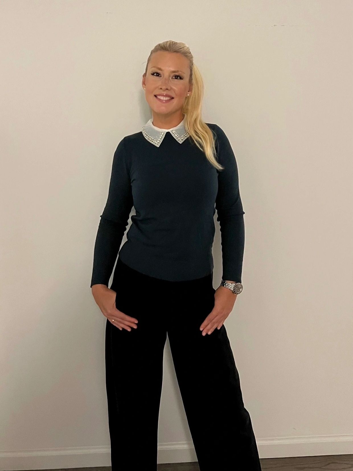Woman in black outfit with a decorative collar, smiling against a plain wall.