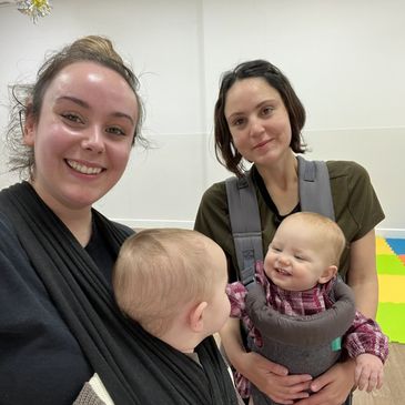 Two women each holding a baby, smiling indoors.