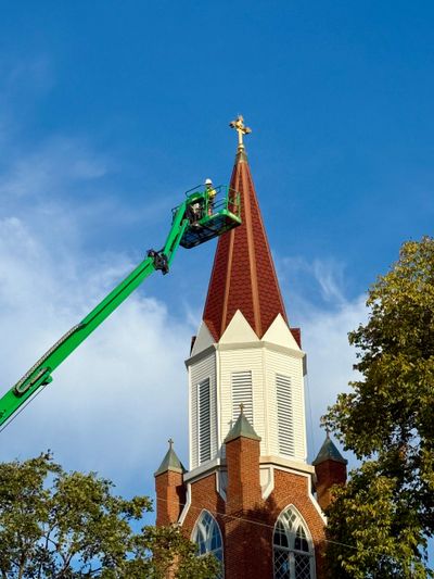 Worker on a lift repairing a church steeple cross under a clear blue sky.