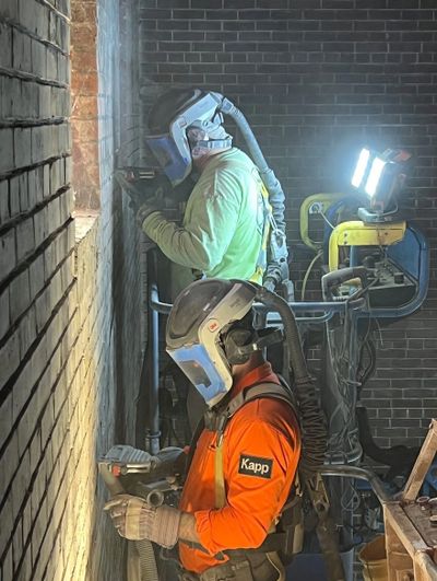 Two workers in protective gear using tools on a brick wall indoors.