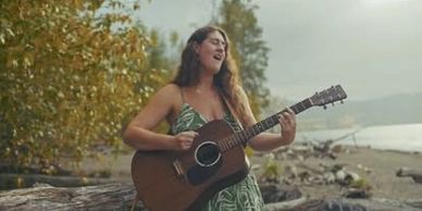 Woman playing guitar and singing outdoors near a beach.