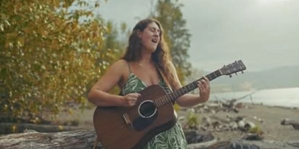 Woman playing guitar and singing outdoors near a beach.