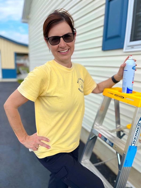 Woman in sunglasses holding spray can, standing by a ladder outside a house.