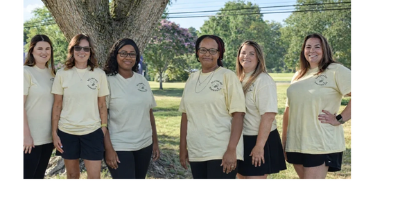 Group of six women wearing matching light yellow t-shirts outdoors.