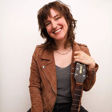 Smiling woman holding a guitar, wearing a brown jacket.