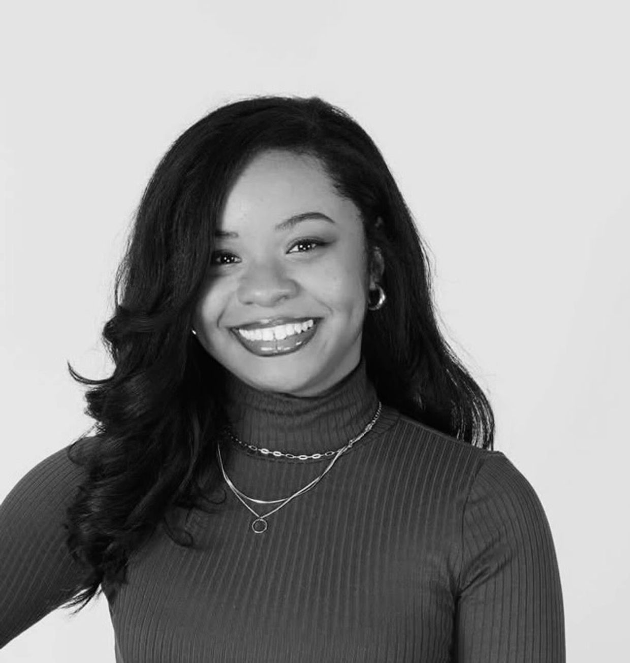 Smiling woman with wavy hair in a ribbed turtleneck and layered necklaces.