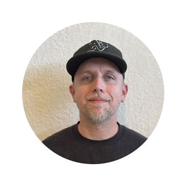 Man wearing a black baseball cap and black shirt, smiling slightly.