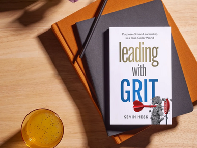 Book titled 'Leading with Grit' on stacked notebooks with a pencil and a drink nearby.