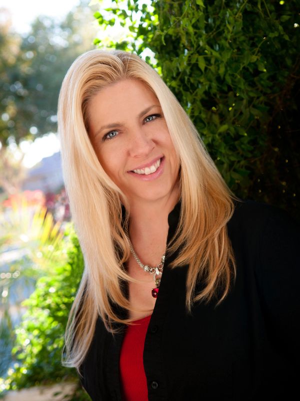 Smiling blonde woman outdoors with greenery in the background.