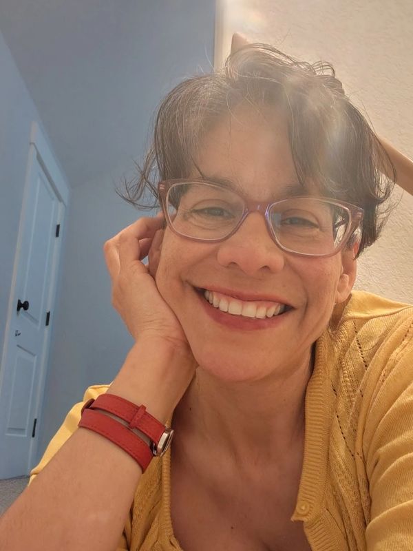 A smiling woman with glasses and a red watch strap, wearing a yellow top.