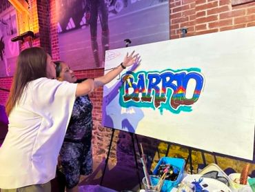 Two women painting graffiti art that says 'BARRIO.'