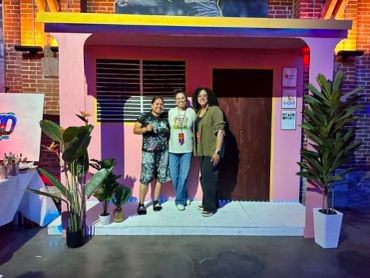 Three people posing in front of a colorful structure decorated with plants.