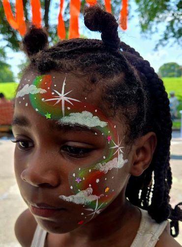 Child with colorful face paint of stars, clouds, and rainbow swirls.